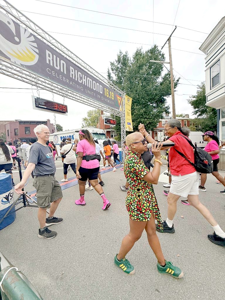 Kelli Lemon dancing with a microphone under the Run Richmond 16.19 arch at the start of the race