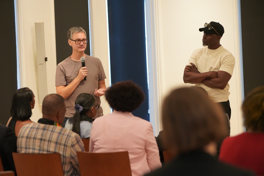 Max Plank speaking into a microphone beside Djimon Hounsou at the Black History Museum & Cultural Center of Virginia